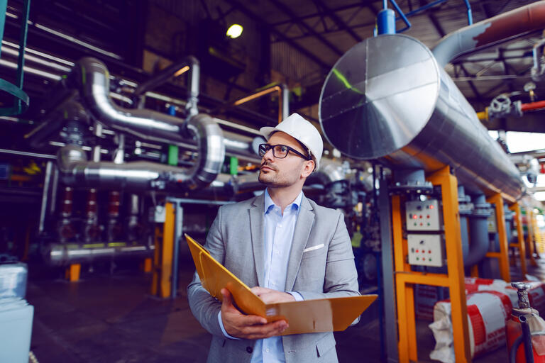 Handsome caucasian businessman in suit and with protective helmet on head holding folder with documents while standing in power plant.