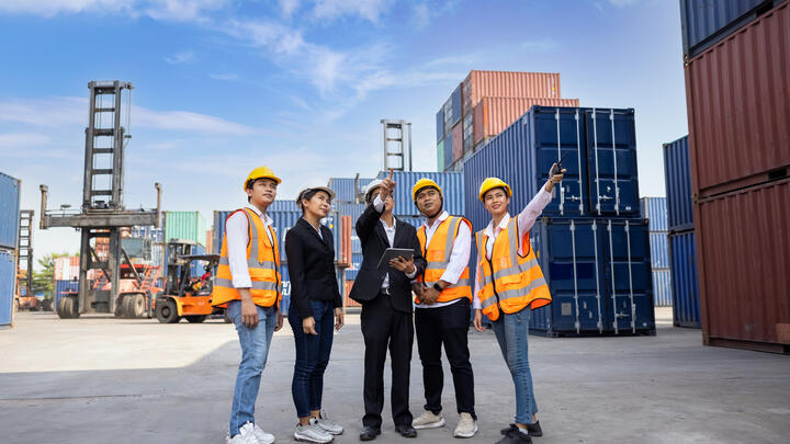 Team of logistic engineer man and woman wearing helmets with tablet holding hand and pointing finger standing outside containers area warehouse